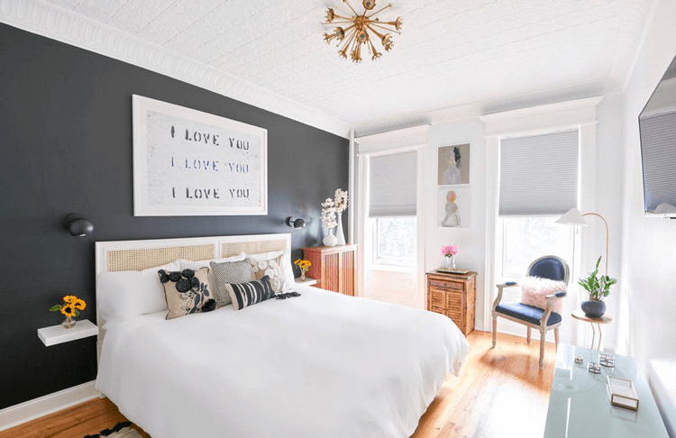 charcoal black accent wall, white bedroom, eclectic bedroom, honey oak flooring, oak flooring, white bedding, navy chair, pink sheep skin pillow, starburst light fixture, honeycomb shades